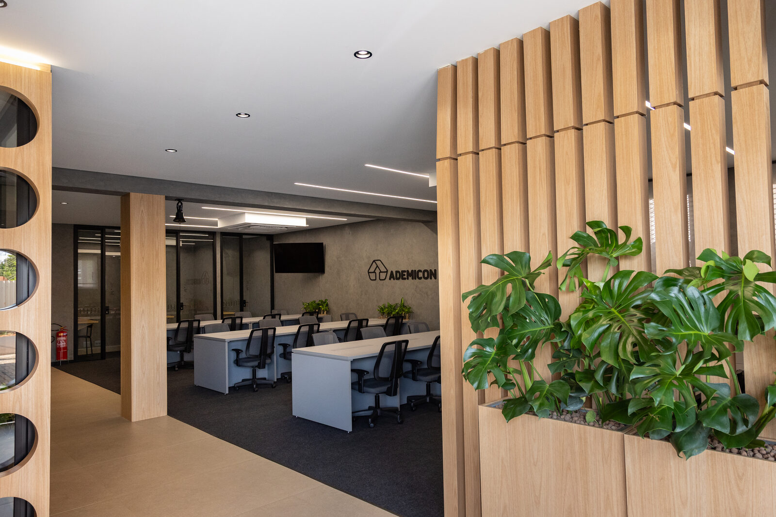 Hall do escritório no Alto da Boa Vista, em Ribeirão Preto. Ripado de madeira, plantas tropicais e logo Ademicon em alto-relevo na parede de cimento queimado.