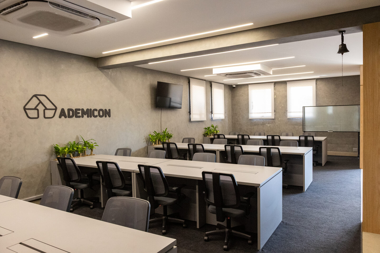Sala de treinamento e operação comercial com logo Ademicon em alto-relevo na parede de cimento queimado e estações para a equipe.