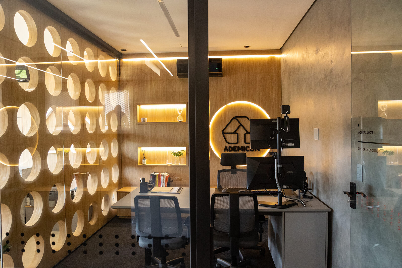 Sala de atendimento executivo com painel de madeira de círculos vazados, iluminação dourada e logo Ademicon circular em LED.